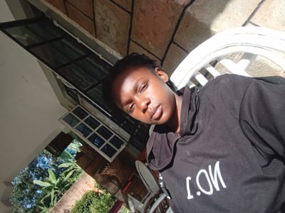 A young African woman with braided hair is taking a selfie outdoors. She is wearing a black hooded sweatshirt with the white text "I.ON" on it. The background suggests a rustic, possibly rural or semi-rural setting, with a stone-tiled wall and a partially visible white chair. There is also greenery and a window with blue panes visible. The lighting indicates it is daytime, with sunlight casting shadows. The image is taken from a high angle, looking down at the subject. The location is identified as Mahiakalo ward, Kenya.