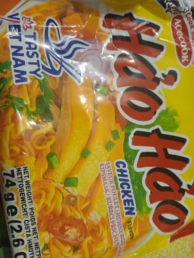 The image is a close-up of a package of Hao Hao instant noodles, specifically the chicken flavor. The package is predominantly yellow with bold red lettering that spells out "Hao Hao". Below the brand name, "CHICKEN" is written in blue, followed by "Flavour" in yellow. Text in multiple languages, including French, German, and Czech, appears to describe the flavor.

The foreground of the package features a stylized image of cooked chicken slices, noodles, green onions, and a sprig of parsley. To the left, a blue and white logo contains stylized chopsticks and the words "TASTY VIET NAM" in blue lettering. The background shows a blurred green leafy vegetable, possibly lettuce. The lighting suggests it is an indoor shot, likely in a store or home. The overall impression is that of a food product advertisement, aiming to be visually appealing and informative about the contents. There is no discernible time of day, weather, or specific location cues beyond the product's origin implied by "VIET NAM". The emotions conveyed are neutral, focusing on the product presentation.