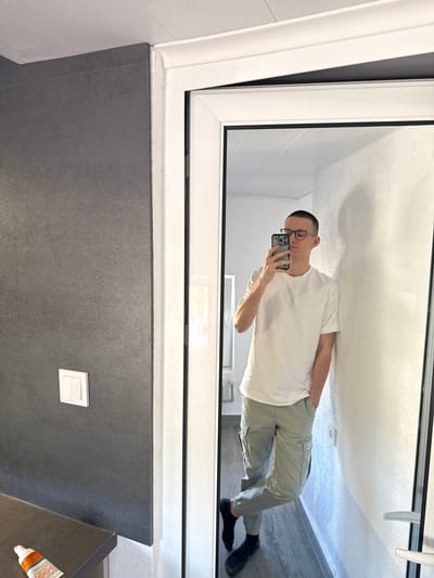 A man takes a mirror selfie in what appears to be a bathroom or hallway. He is wearing a white t-shirt, light green cargo pants, and black socks. He has short brown hair and is wearing glasses. He is leaning against a white wall, with his right hand in his pocket. In the reflection, a grey tiled wall is visible to the left, with a white light switch. A tube of what appears to be toothpaste or lotion is on a dark countertop in the bottom left corner of the image. The floor is wooden. The man is standing in front of a door or mirror, which reflects the opposite wall, also white and textured. The ceiling appears to be made of white panels. The overall environment suggests an indoor, possibly residential setting. The location in the city of Momchilovtsi, Bulgaria, is not directly indicated by any visual cues in the image.
