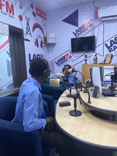The image depicts a radio studio at LASU Radio 95.7 FM, a campus radio station situated in Ijanikin, Nigeria. The studio is equipped with a sound mixer, microphones, computer keyboard, and a television mounted on the wall. A banner featuring the radio station's logo, name, and the phrase "Solely Unique Campus Radio Station" is visible on the wall. Two individuals are present in the studio, seated behind the table, wearing headsets and positioned at microphones. The environment appears to be a professional broadcast setting.