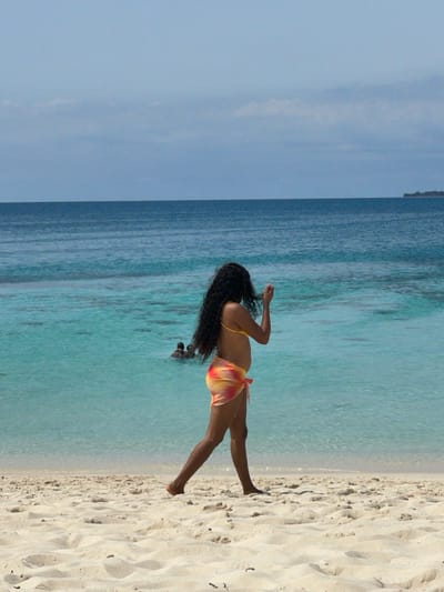 A pristine sandy beach environment is depicted in Zanzibar, Tanzania, under a bright sky. In the foreground, a person with long, dark, wavy hair walks barefoot from left to right. This individual is wearing a yellow bikini top and a translucent sarong with a red, orange, and yellow tie-dye pattern, tied at the waist. Their right hand is raised to the head, seemingly adjusting their hair. In the midground, the water is clear and turquoise, gradually deepening to a darker blue towards the horizon. Two indistinct human figures are partially submerged in the turquoise water on the left side of the midground. The far background shows the ocean meeting a light blue sky with minimal cloud cover, and a faint, hazy outline of a small island or landmass is visible on the far right horizon.