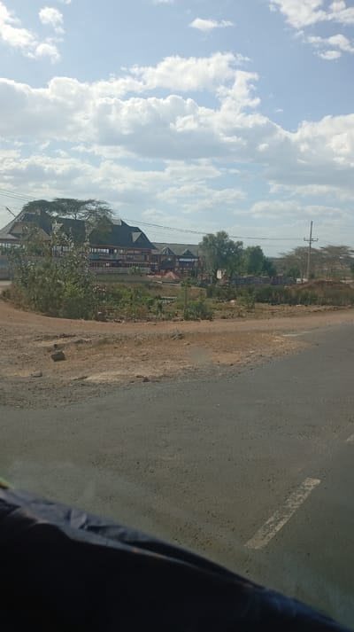 A roadside view shows a building complex with a dark roof and visible signage amidst greenery. The building is likely a hotel or restaurant. A dirt area and a paved road are in the foreground. Above, a bright sky features many fluffy white clouds. The photograph is taken from a vehicle, with a dark fabric or object visible at the bottom.
