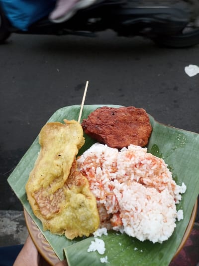 On a banana leaf, a dish presents fried tempeh with a toothpick, a square piece of reddish-brown meat, and rice mixed with reddish flakes. The dish rests on a patterned plate. A blurry motorcycle and a piece of trash are visible in the background, along with a bit of someone's lap or knee.