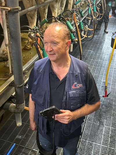In Wilhelminaoord, Netherlands, a middle-aged Caucasian man with fair skin and thinning, light brown hair stands in what appears to be a modern dairy farm. He is wearing a dark blue vest over a dark blue collared shirt, blue jeans, and a black lanyard around his neck. The vest has a red and white logo on the left chest area. In his right hand, he holds a dark-colored electronic device, possibly a tablet or scanner, which is connected to the lanyard.

The man is positioned in the foreground, facing slightly to his left and looking upwards and to his left. The background reveals a milking parlor setup. Several cows are visible, standing in stalls with their udders connected to milking equipment. The milking apparatus consists of various hoses, tubes, and cups, predominantly in green and orange colors. The floor of the parlor is a metal grate, with a channel of murky liquid visible between the stalls. To the right of the man, a yellow hose is attached to a red spigot on the wall. The overall environment suggests a working farm facility focused on dairy production.