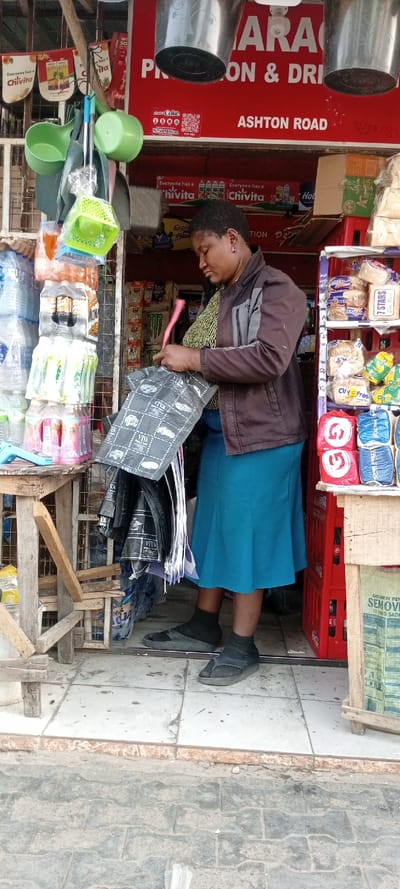 A woman stands in the entrance of a shop, arranging black plastic bags. She's wearing a brown jacket, a blue skirt, and black flip-flops with socks. Behind her, the shop displays a variety of items including water bottles, bread, and Chivita products. A red sign above reads "GARAGE, PROVISION & DRI," with "ASHTON ROAD" below. Metal buckets hang from the ceiling. The shop appears to be a small, local business.