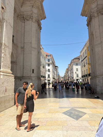 A sunny, wide, cobblestone street in Lisbon, Portugal, is shown from ground level. Two people, a man and a woman, stand in the foreground, facing away from the camera. The man wears a dark t-shirt and khaki shorts, and the woman wears a black dress. They are standing near a large, stone archway.
In the background, the street is lined with multi-story buildings with balconies and windows. A crowd of people walks along the street, some looking at shops, others conversing. The sky is clear and blue. The scene appears to be a popular tourist area, with many people enjoying the warm weather.