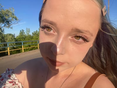 The media file is an upside-down, close-up selfie of a young woman. She is outdoors, with a bright blue sky and green trees visible in the background. A yellow and blue railing runs horizontally across the image, suggesting a bridge or elevated walkway. The sun is shining brightly, casting a distinct shadow across the woman's face. Her skin appears shiny, possibly from sweat or makeup. She is wearing a necklace and a top with a floral pattern. Her eyes are green and her eyelashes are long and dark. The overall impression is that she is enjoying a sunny day outdoors in Nova Praha, Ukraine. No text is visible in the image.