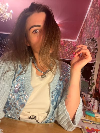 In the city of Krasnoe-na-Volge, Russia, a medium shot captures a woman seated at a wooden table. She is wearing a light blue, knitted cardigan adorned with large, iridescent blue and clear embellishments, over a white top. A beaded necklace with a sun-shaped pendant rests on her chest. Her right arm is bent, with her hand near her face, revealing a tattoo on her forearm and bright pink nail polish. Her dark hair partially obscures her face, with strands falling across her left eye. The background is a domestic interior featuring floral wallpaper and a pink ceiling with a chandelier. Various objects, including bottles, brushes, and papers, are visible on the table and in the background. The lighting suggests an indoor, possibly nighttime setting.