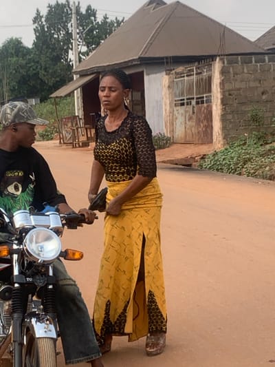 An outdoor scene in Aguleri, Nigeria, depicts two individuals on an unpaved, reddish-brown dirt road. On the left, a man is seated on a motorcycle, partially visible. He wears a camouflage-patterned cap, a black t-shirt with a green design, and blue jeans. His left hand rests on the motorcycle's chrome handlebar, near a white container. The motorcycle features a large circular headlight and orange turn signals. To the right of the motorcycle, a woman stands facing forward, her head slightly turned to her left. She wears a black lace top over a yellow patterned garment, a long yellow skirt with black patterns at the hem and a high slit on the left side, and platform sandals. Her right hand holds a dark-colored handgun, positioned against her lower abdomen. In the background, two buildings are visible: one with a dark corrugated metal roof and a light-colored facade, and another with a brick base and a rusty metal gate. Scattered vegetation, including trees and bushes, is present behind the buildings. The sky is overcast.