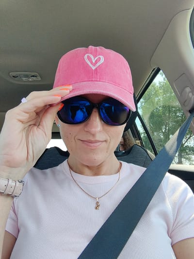 The image is a selfie taken by a woman inside a car. She is wearing a pink baseball cap with a white heart embroidered on it, dark sunglasses, and a light pink ribbed t-shirt. She is also wearing a delicate gold necklace with a cat pendant and a light pink watch on her left wrist. Her right hand is holding the brim of her hat. The car's interior is visible, including the grey headliner and a portion of the dark grey seatbelt. Outside the car window, trees and a bright sky are visible. The scene does not provide any specific visual cues to determine its location within Дзяржынскі сельскі Савет, Belarus.