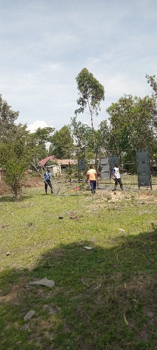 The image depicts an outdoor, daytime scene in Kisumu, Kenya, under a partly cloudy sky. The foreground and mid-ground consist of an uneven grassy area with patches of bare earth and sparse vegetation.

Three adult males are observed in the mid-ground. To the left, an individual wearing a light blue long-sleeved shirt and dark trousers is bending over, manipulating or interacting with a section of a silver-colored metal truss structure that lies horizontally on the ground. In the center, a second individual, dressed in an orange t-shirt and dark trousers, is walking towards the right side of the frame. To the right, a third individual, wearing a white sleeveless vest with blue accents and dark trousers, stands upright, facing towards the left.

Behind the central and right figures, a setup of three dark-colored rectangular structures is visible. These structures appear to be professional audio speaker stacks or frames designed for speaker arrays; the rightmost stack is largely upright and composed of multiple individual units.

The background features dense green foliage and numerous trees. Beyond the immediate tree line, partially obscured by vegetation, are several buildings with reddish-brown corrugated metal roofs, indicating a settlement or residential area.