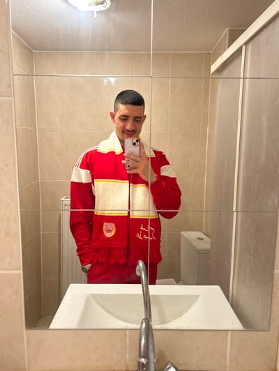 A young man is depicted in a mirror selfie, smiling broadly with his eyes partially closed, suggesting a cheerful or amused expression. He has short, dark hair and is wearing a red tracksuit top with prominent white stripes running down the sleeves. Around his neck, he sports a red and white striped scarf, complete with fringes at both ends. One end of the scarf features a golden or yellow embroidered crest, possibly depicting a wreath or circular emblem with an internal design. The other end of the scarf has white embroidered Arabic script or stylized text. He is holding a smartphone, which appears to be an iPhone with a light purple or pink case, in his left hand to capture the image.

The setting is a bathroom, as indicated by the presence of a white ceramic sink with a chrome faucet visible to the right of the man. The sink bowl shows some visible watermarks or residue. The walls are tiled with light beige or cream-colored square tiles. The mirror itself, where the reflection is captured, consists of multiple panels evident by faint vertical and horizontal dividing lines, and it appears slightly smudged or dusty in areas. A light fixture is visible on the wall to the left of the man's reflection, providing illumination. In the background reflection, behind where the man stands, a white object resembling a radiator or part of a toilet cistern can be seen. The image is taken indoors, likely under artificial lighting.