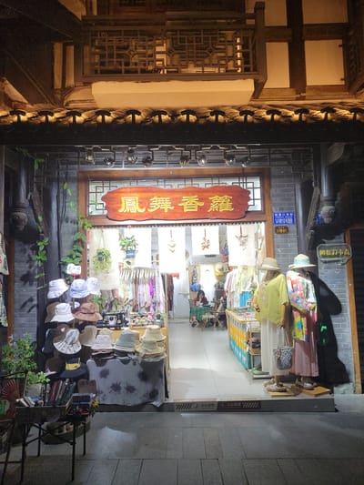 Here is a description of the image:

This nighttime image shows a shop front, likely in the Shapingba District of China, based on the signage. The shop appears to sell clothing and accessories, with mannequins near the entrance displaying various outfits and hats. A prominent vertical sign in Chinese characters hangs above the doorway.

The shop's exterior has a traditional aesthetic, with detailed woodwork. Lantern-like fixtures illuminate the entrance. Various hats and other items are displayed outside the shop on tables and stands. Plants are growing along the shop's façade. The street appears paved with stone or concrete slabs.