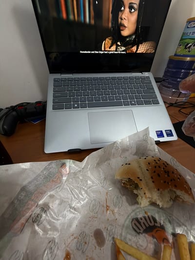 The image displays an indoor scene in Jiangning District, China, focusing on a casual meal and entertainment setup. A silver laptop, open on a brown desk or table, serves as a central element. The laptop features "Intel Core Ultra 7" and "Intel Arc Graphics" stickers prominently displayed on its palm rest. Its screen is either displaying content or connected to a larger television screen positioned vertically to the left of the laptop.
The television screen shows a close-up of a woman with dark hair and striking eye makeup, presumably from a show or movie. Subtitles are visible at the bottom of the screen, reading: "Homelander and Stan Edgar had a good heart-to-heart," which is a line from the TV series *The Boys*.
To the right of the laptop, a partially eaten burger with sesame and black sesame seeds on its bun, along with several french fries, rests on grease-stained white wrapping paper. The paper features a recurring logo of a stylized burger, indicating fast-food packaging. A dark logo with text, possibly "BURU," is also visible through the wrapping paper near the fries.
In the upper background, stacked blue and white containers are visible, one of which clearly shows the Chinese characters "乳牛奶" (Rǔ niúnǎi), meaning "Milk," confirming a local context. A black and red gaming controller is partially visible beneath the laptop, suggesting additional leisure activities. The overall scene depicts a relaxed setting, combining eating a fast-food meal with watching media.