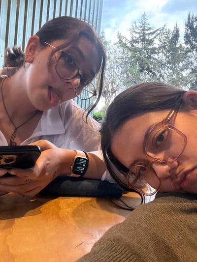 Two young women pose for a selfie. The woman on the left wears eyeglasses and has her tongue sticking out slightly. She is dressed in a white shirt and has a necklace. Her left hand holds a dark-colored smartphone, and she wears a gold-colored Apple Watch on her left wrist. The woman on the right wears oversized eyeglasses and puckers her lips as if giving a kiss. She is wearing a grey sweater. They appear to be sitting at an outdoor table, as suggested by the wooden surface and corrugated metal wall in the background. The sky and trees are visible above them. The photo was taken outdoors in the city of San Andrés Cholula, Mexico.