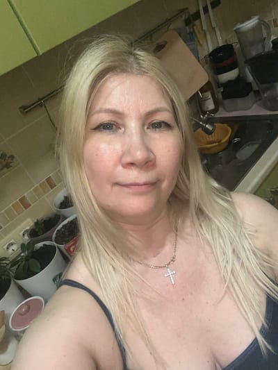 A woman with blonde hair is taking a selfie in a kitchen. She is wearing a dark blue top and a necklace with a cross pendant. In the background, there are kitchen cabinets, shelves with dishes, a sink, and several potted plants. The overall setting appears to be domestic. The image does not provide any information about the city of Shushary, Russia.