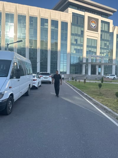 In Manas, Kyrgyzstan, a man in dark clothing walks on an asphalt road in front of a modern, multi-story building with large glass windows. A white Mercedes-Benz Sprinter van is parked to the left, and several other cars are visible in a row behind it. The building features a prominent emblem on its facade. A well-maintained lawn with scattered trees and a bench lines the area in front of the building. The sky is clear and blue, indicating daytime.