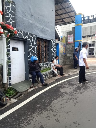 A gray building with a black and white rock pattern sits next to a street. A blue-helmeted individual is seated on the building's ledge, next to a man and a child. Further down, two men are talking, and another man stands in the street. A building with "SM" on top is visible in the background, along with a blue entrance gate with a warning sign. A strip of green plants grows at the base of the building.