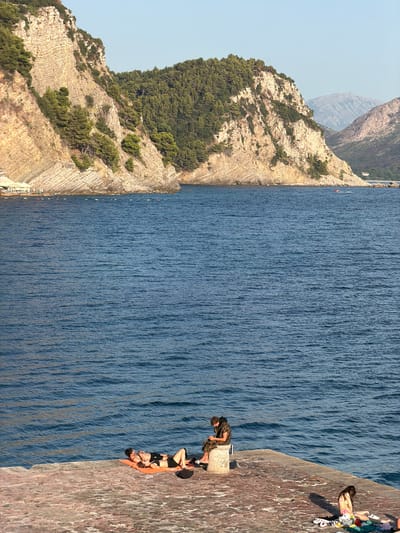 In Petrovac centar, Montenegro, a coastal scene unfolds under a clear sky. The foreground features a stone pier where two individuals recline on an orange towel, one appearing to be a male with dark hair wearing black shorts and a bare chest, the other also wearing dark shorts. Nearby, a second individual, possibly male, sits on a stone pillar, engaged with a mobile device, dressed in a patterned hooded jacket and shorts. Further to the right, another person, appearing female, sits on a towel with colorful items, wearing a pink bikini. The pier overlooks a calm, deep blue body of water. In the background, a dramatic, layered cliff face rises, densely covered with dark green pine trees. The cliff slopes down to the sea, where a small section of a beach with white umbrellas and lounge chairs is visible. In the distance, hazy mountains form the backdrop, with a small white building nestled on the coastline. A small red and white motorboat is visible in the water near the distant shoreline.