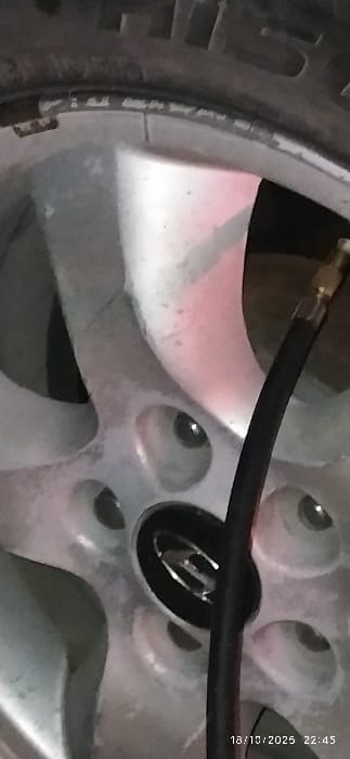 This image captures a close-up, vertical view of a silver-colored alloy vehicle wheel and the upper portion of a black rubber tire. A black air hose, equipped with a brass-colored metallic connector, is inserted into the tire's valve stem, indicating tire inflation or pressure assessment. The multi-spoke alloy wheel exhibits minor superficial dirt and wear. A black center cap featuring a reflective, oval-shaped emblem, which is not clearly identifiable, is present. The tire's tread pattern and sidewall are partially visible at the top of the frame. The background is dark and out of focus, with a faint reddish light reflecting on parts of the wheel structure. No individuals are depicted in the scene. A timestamp reading "18/10/2025 22:45" is visible in the lower right corner. The scene is located in Al-Kiswa, Syria.