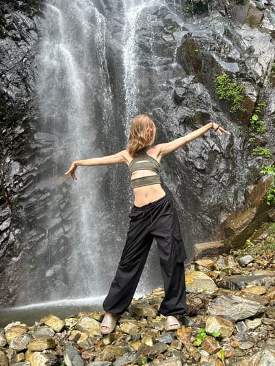The image depicts a young person posing in front of a waterfall in Tu Le, Vietnam. The subject, a female with blonde, wavy, shoulder-length hair, is positioned centrally, facing away from the camera and slightly to the left. She wears an olive-green crop top with horizontal cut-outs across the midriff and loose-fitting black cargo pants. Light beige open-toed slide sandals cover her feet. Both arms are extended horizontally outwards, with hands slightly cupped downwards; her right arm is slightly higher and more extended than her left, adorned with multiple dark and one pink bracelet near the wrist. The waterfall behind her features a significant volume of water cascading down a dark, jagged rock face, forming a white, frothy pool at the base. Patches of green vegetation are visible on the wet rocks surrounding the waterfall. The foreground consists of an uneven terrain composed of various sizes of wet, multi-colored rocks (grey, brown, yellow), with sparse green plant growth scattered among them. The scene is illuminated by natural daylight, suggesting an outdoor environment.