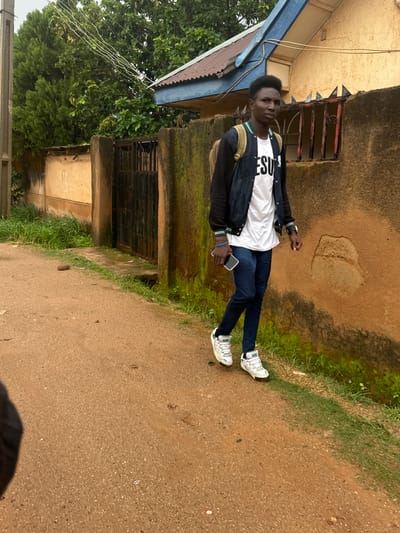 The image shows a young man standing outdoors in what appears to be a residential area. He is wearing a white t-shirt with black text that reads "JESU", dark blue jeans, and white sneakers. He is also wearing a dark jacket with green and white accents. He has a backpack slung over one shoulder and is holding a smartphone in his right hand.
The setting is characterized by a building with a yellow-brown textured wall and a blue corrugated metal roof extension. There is a barred window on the building, and moss is visible on parts of the wall. To the left of the building, there is lush green vegetation, including trees and grass. The ground is a reddish-brown, gritty surface, suggesting it could be a dirt road or pathway. A patch of grass lines the edge of the pathway.
The image is taken from a slightly elevated perspective, looking down at the man. The lighting suggests it is daytime, and the overall atmosphere appears casual and everyday. The man's posture and gaze do not convey strong emotions, but he appears relaxed.
Visible text: "JESU" on the t-shirt.
Location context: Gwong, Nigeria.