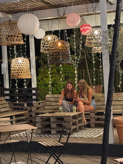The image shows an outdoor seating area at night in Cavriglia, Italy. Two young women are seated on a bench made of wooden pallets, facing a low table also constructed from pallets. The woman on the left has long brown hair and is wearing a black top and a striped skirt. The woman on the right has long blonde hair and is wearing a black top and denim shorts. Both women are looking at their phones. On the table between them are two glasses with drinks and ice.
The seating area is decorated with numerous hanging lanterns, some made of woven materials and others of paper in various colors including white, pink, and red. String lights are draped around the seating area, providing illumination. The furniture is rustic, composed of repurposed wooden pallets. In the background, there is lush green foliage and a metal railing. A large terracotta pot is visible on the right side of the image. The overall atmosphere suggests a casual outdoor gathering space.