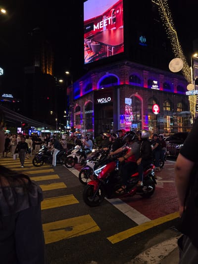 The image depicts a bustling street scene at night in Kuala Lumpur, Malaysia, likely within the Bukit Bintang area, known for its vibrant nightlife and shopping districts. The foreground is dominated by a busy pedestrian crossing with bright yellow stripes on dark asphalt. Several people, some on motorcycles, are waiting at the crossing.
In the mid-ground, a group of motorcyclists and pedestrians are visible. Many of the motorcyclists are wearing helmets and casual attire. Behind them, buildings are illuminated with neon lights and large digital billboards. One prominent billboard displays the word "MEET" with an image of a modern interior. Another building features the "WOLO" logo. The architecture suggests a mix of modern and older structures, with brickwork and arched windows visible on one building.
The background shows more city lights, including traffic lights and street lamps, indicating a busy urban environment. Decorative string lights are also visible, possibly for festive purposes. The overall atmosphere is lively and energetic, characteristic of a popular entertainment district at night. The presence of the "WOLO" logo strongly suggests the scene is located at or near the WOLO Bukit Bintang hotel.