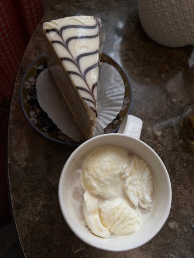 A slice of vanilla cake with dark chocolate drizzle, presented on a decorative plate, sits next to a white mug filled with melted vanilla ice cream. The scene is captured from a top-down perspective, with the dark, speckled surface of a table as the backdrop. The lighting suggests an indoor setting, possibly a cafe, and no individuals are visible. The location of Petropavlovsk-Kamchatsky, Russia, is not ascertainable from the visual information provided in the image.