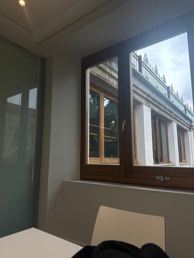 A view from inside a building in Segovia, Spain (latitude: 40.953, longitude: -4.119). The scene is captured through a large window, positioned at the top of the room, looking out at the exterior of another building. The exterior building features a white facade with multiple windows and a black metal fence at the top. The window is framed in wood and is open. The interior of the room is finished with white walls and a light grey tiled floor. The only light source is a small spotlight in the ceiling. The window is reflecting the surrounding environment, creating a blurred image of the exterior building and the sky. The overall impression is of a bright, airy room with a view of a modern building.