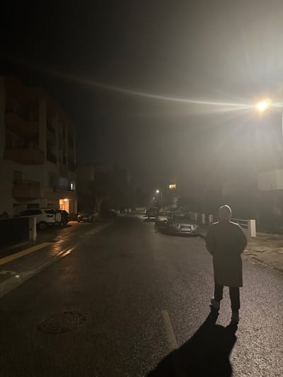 The image captures a nocturnal scene on a wet street in Engomi, Nicosia, Cyprus.

**Subjects:**
A single adult figure, largely silhouetted, stands on the left side of the wet asphalt road. The person is wearing a light-colored coat or jacket and dark trousers, with a light-colored head covering or hair. Their long shadow stretches across the road to the right. On the left, a bright street light emits a strong glare and lens flare, illuminating the immediate area. Several cars are parked along the street, including a dark-colored sedan or hatchback visible in the mid-ground. At least one white motorbike or scooter is parked closer to the foreground. Multi-story buildings with balconies line both sides of the street, some displaying interior lights or appearing partially constructed with visible scaffolding. Dark, indistinct vegetation is visible between some buildings. A manhole cover is present on the road surface.

**Activity or event taking place:**
A person is standing on a street at night, seemingly observing their surroundings. No dynamic activity is depicted.

**Notable details:**
The time of day is clearly nighttime, characterized by the dark sky and reliance on artificial light sources. The street appears wet and reflective, suggesting recent rain or damp conditions. The strong light diffusion from the street light could indicate atmospheric moisture like mist or fog, or simply camera lens flare. The architecture of the buildings and the style of vehicles suggest a Mediterranean or European urban setting, consistent with the provided location context of Cyprus. The scene conveys a somewhat solitary or contemplative mood due to the lone figure in the dark, wet environment.

**Visible text:**
Faint characters are visible on the license plate of the parked car, including what appears to be "52" followed by indistinct letters.