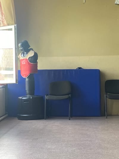 The image shows a room that appears to be a martial arts training area or gym. In the foreground, there's a punching bag with a black base and a corrugated black middle section. A red and white chest protector, possibly for martial arts, is positioned on top of the punching bag. To the left of the punching bag, there's a white framed object, which might be a mirror or a display case.
In the background, there's a large blue mat covering a significant portion of the floor. Behind the blue mat, there are two black chairs with metal frames. The walls are a light, neutral color, and the floor appears to be carpeted. The lighting in the room is bright, suggesting it's daytime, but there are no windows visible to indicate the exact time or weather conditions. The overall impression is one of a functional training space, likely indoors. There is no visible text in the image. The scene is static, with no apparent activity taking place at the moment the photo was taken.