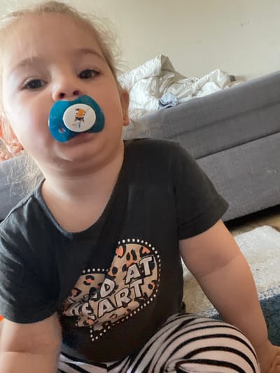 A child is lying on their side, with a blue pacifier in their mouth. The child is wearing a black t-shirt with a beige and black graphic print and striped leggings. The child appears to be lying on a couch or bed with bedding or a blanket. The location is Senden, Germany, but nothing in the image can confirm this.