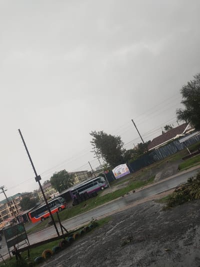 The image captures an outdoor scene in Ngong ward, Kenya, under a predominantly grey, overcast sky. The ground exhibits wet asphalt and dirt patches, indicating recent precipitation. In the middle ground, two buses are stationary: one primarily orange and red, and another purple with lighter grey sections. Behind these vehicles, multiple multi-story buildings are visible. To the right, a white sign with colorful text displays "HARMONY HILL SCHOOL," positioned next to a dark corrugated fence. Further right, a building with a red-tiled roof is partially obscured by the fence. Utility poles and overhead wires crisscross the upper and middle sections of the frame. The left foreground features a tall, slender dark pole with regularly spaced horizontal protrusions and a row of approximately ten horizontally placed tires, painted in varied colors. Patches of green foliage, including trees and bushes, are distributed across the scene. No individuals are visible.