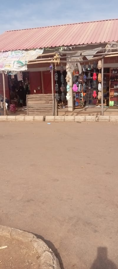 The image displays a street scene in Kasuwan Magani, Nigeria, under a clear blue sky. A single-story building with a red corrugated metal roof occupies the upper half of the frame. Below the roof, a makeshift awning, constructed from patterned sacks or tarps, is supported by rough wooden poles and beams.
The area beneath the awning features two visible retail sections. On the left, an open shop entrance reveals a dark, indistinct interior, fronted by a low wooden slat barrier or counter. To the right, an extensive collection of footwear, predominantly sandals and flip-flops, is displayed. Numerous pairs in various colors (black, white, pink, blue, red, brown, tan, green) are suspended from strings hanging from the overhead structure. Additional pairs are neatly arranged on light-colored wooden shelves within an open display to the right.
The foreground comprises an unpaved, light brown earth road exhibiting scattered debris and faint tire marks. A low, grey concrete curb separates the road from the raised storefront area. A small blue object is visible on the road near the curb, slightly to the left of the center. The shadow of an individual is cast on the lower right portion of the unpaved road.