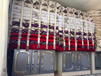 A vertically oriented, medium shot captures a stacked collection of pillows and bedding in Sovet, Kyrgyzstan, filling a significant portion of the frame. The items are organized in neat rows, with some featuring intricate patterns of purple, red, and geometric designs, while others are plain white with floral motifs. Decorative tassels and woven elements hang between the rows, adding a handcrafted touch. Below the bedding, two dark gray cabinets with ornate gold trim and ribbed detailing are visible. The left cabinet features a door and a narrow panel, while the right cabinet has two narrow panels. A portion of a white lace curtain hangs from the left edge of the frame, and a dark doorway is partially visible behind it. The background is a plain, light-colored wall. The lighting is even, suggesting an indoor setting, possibly a room dedicated to storage or display of traditional textiles.