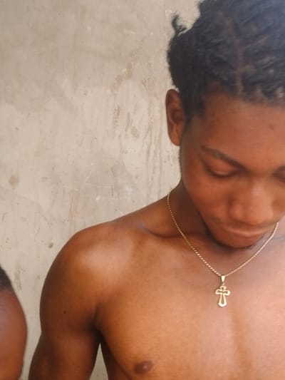 The image depicts a close-up of a bare-chested individual with medium-dark skin, observed in Enugwu-Ukwu, Nigeria. The subject's head is tilted downwards and to the right, with eyes directed away from the viewer. Dark hair is styled in braids or twists, pulled back from the face. A thin, gold-colored chain with a stylized cross pendant hangs around the subject's neck, resting on the upper chest. The left nipple is visible. A portion of a second individual's dark-skinned shoulder or upper arm is partially visible on the far left side of the frame, suggesting proximity. The background consists of a textured, light-colored wall exhibiting discolorations and streaks, consistent with weathering.