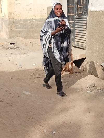 The image depicts an adult female walking outdoors on an unpaved dirt path in Gwagwarwa, Nigeria. The subject, with dark skin, wears a black and white tie-dye patterned head covering and drape, dark pinstriped trousers, and dark open-toed sandals. She holds a dark-colored smartphone in both hands near her upper chest/lower face, displaying a wide smile with an open mouth. A small reddish-brown animal, possibly a goat, is partially visible to her right, near a concrete structure. The background includes a dark, paneled door on the right and a light-colored, weathered wall with faded markings on the left. The ground is composed of light brown dirt/sand with minor scattered debris. The scene is illuminated by strong overhead sunlight, creating distinct shadows.