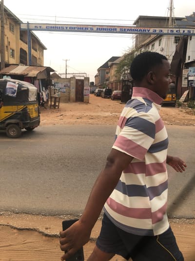 The image captures an outdoor urban or semi-urban scene in Akpaka Forest Reser, Nigeria, under an overcast or hazy sky. In the foreground, a dark-skinned male subject, with short dark hair, is walking from right to left, mid-stride. He is dressed in a short-sleeved collared shirt featuring horizontal stripes of white, light pink, and light blue, paired with dark shorts. A black mobile phone is held in his left hand, partially obscured by his leg.
In the midground, an overhead blue archway sign is prominent, displaying the text "DIOCESE ON THE NIGER (ANGLICAN COMMUNION) ST. CHRISTOPHER'S JUNIOR SEMINARY 3-3. (S.C.J.S.)". To the left of the paved road, a yellow and black motorized tricycle is partially visible, parked near a makeshift market stall with a corrugated metal roof and various items of clothing displayed. A wooden utility pole stands near the center of the image.
Beyond an open brown metal gate on the right, a dirt road extends into the background, where multiple vehicles are parked or visible. Multi-story buildings line both sides of the street, including a light-colored structure on the far left and a building with visible scaffolding on the right, behind the archway sign. The immediate ground consists of an asphalt road with a dirt shoulder to the right.
