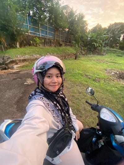A woman is sitting on a blue and black motorbike. She is wearing a light pink helmet with a reflective visor, a black and white patterned headscarf, and a light-colored long-sleeved shirt. The woman is smiling and looking at the camera. She is positioned in the center of the frame, and the motorbike's handlebars and front are visible to her right. A black bag is attached to the motorbike's front.
The setting appears to be outdoors, with lush green grass and trees in the background. The sky is visible in the upper left corner, suggesting it is either early morning or late afternoon, with a warm, hazy light. There is a fence or structure made of metal bars and blue material visible behind some trees on the left. The ground where the motorbike is parked is a mix of dirt and asphalt.
The image is taken in portrait orientation and appears to be a selfie. The overall mood of the image is cheerful and casual. No visible text is present in the image. The context is Banjarbaru, Indonesia.