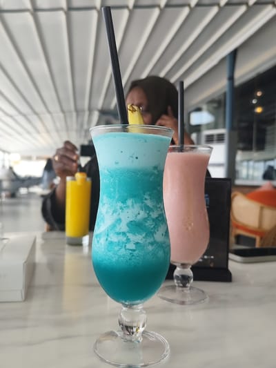 The photograph captures a moment at an eatery in Jeddah, Saudi Arabia. A turquoise-colored, iced beverage with a pineapple garnish and black straw stands prominently in the foreground. Beside it is a pink-colored drink in a similar glass. A black straw is inserted into the drink as well. A woman with a dark head covering is seated in the background, using a mobile phone. Beside her are two other glasses on the table. The setting appears to be indoors, with a light-colored, marble-patterned table surface and a roof structure consisting of parallel slats.