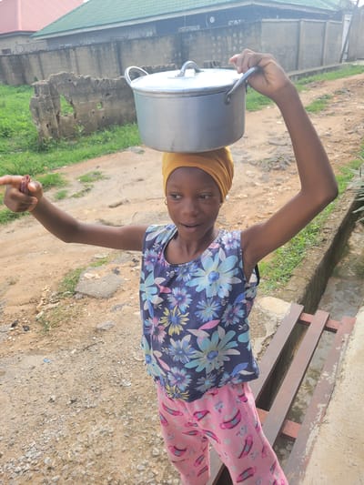 A young woman, likely a teenager, stands on a dirt and gravel surface in what appears to be an outdoor environment. She is wearing a yellow head covering, a floral print sleeveless top, and pink patterned pants. She balances a large, silver cooking pot on her head with one hand. Her other hand holds a small, reddish-brown object. Behind her, there is a dilapidated wall with a building and green vegetation visible. To her right, there is a wooden bench against a light-colored wall. The location is Madalla, Nigeria.