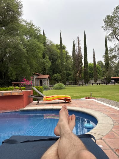 A photograph captures a leisure scene, likely in Cañada de Madero, Mexico, given the landscape features. A pair of bare feet, presumably belonging to a person relaxing on a navy-blue lounge chair, are prominently featured in the foreground, partially submerged in the turquoise blue water of a small, tiled pool. The edge of the pool is bordered by light brown stones. A colorful striped towel hangs on a wooden chair beside the pool. Beyond the pool area, a lush green lawn extends towards a line of tall, thin trees, possibly cypress. Further back, a stone structure with a tiled roof is partially obscured by dense foliage. The sky is overcast.
