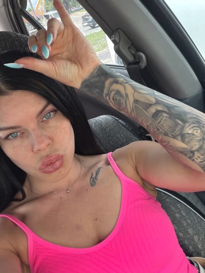 The image shows a woman with long black hair, blue eyes, and freckles. She is wearing a bright pink tank top and has a tattoo on her arm. Her fingernails are painted a light blue color. She is in a car, and the window shows a blurred background of trees and a street. There is a seatbelt visible in the car. The woman is looking at the camera and making a kissing face. She has a small diamond necklace on. There is a tattoo on her chest that says "Fortuna".