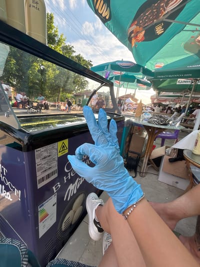 The image depicts an outdoor daytime scene in Kostievo, Bulgaria, likely an open-air cafe or market area. In the immediate foreground, a person's left hand, covered by a light blue disposable glove, forms an "OK" gesture. A silver-colored bracelet with small white stones is visible on the wrist. To the left, a portion of a white sneaker with a black heel tab is present, while to the right, a bare foot with a tattoo on the lower leg is visible.
A dark blue commercial display freezer occupies the left side of the foreground. Its reflective glass top shows an inverted reflection of the background. The freezer features an EU energy label displaying "Class D" and "949 kWh/annum." A warning symbol for flammable R290 refrigerant is present, along with Cyrillic text identifying it as a "JUKA Model: Display freezer R440SL." "Milka" branding is visible on the side, accompanied by an image of round items, possibly pancakes or cookies. Partial text "IT HOCT!" and "lka" is also visible. Above the freezer, partially inverted Cyrillic text "вар а мен" is discernible, possibly from a menu board.
The midground and background reveal a bustling outdoor environment. Multiple people are seated at tables under large commercial umbrellas. These umbrellas display various branding, including "Nestlé," "Milka" (with an image of a chocolate ice cream bar), and "SLUNCHO," along with the Cyrillic text "ПЪРВАТА БЪЛГАРСКА БИРА" (The First Bulgarian Beer). Items on the tables include a stack of ice cream cones, plastic cups, and a zebra-patterned bag. Underneath the tables, boxes and other objects are stored, including one marked "FRAGILISSIMO." Several individuals are walking or standing in the background, and a child's tricycle is situated on the ground. Green trees, a wooden fence, and distant buildings with red-tiled roofs contribute to the backdrop. Overhead, power lines cross a blue sky with scattered white clouds, illuminated by sunlight that suggests late afternoon or early evening.