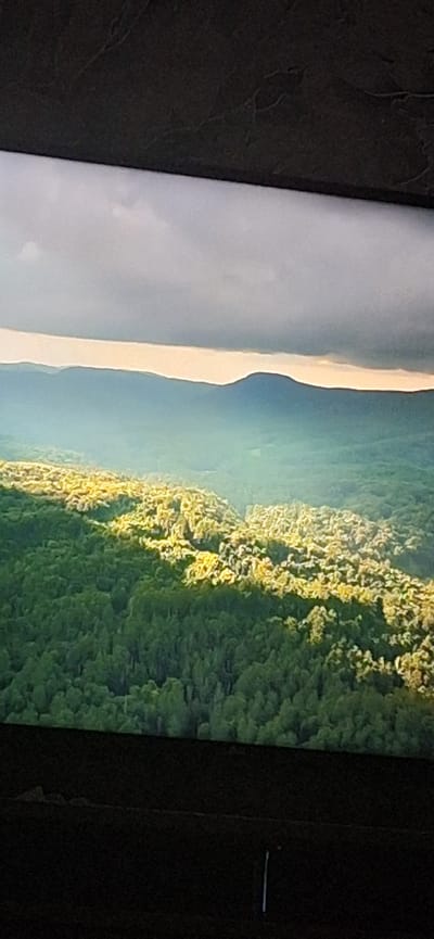 The image displays an aerial view of a densely forested area with rolling hills and distant mountains. Sunlight dramatically illuminates a section of the forest, creating a stark contrast with the shaded areas. A thin, bright band of light is visible between the clouds and the mountains, suggesting a sunrise or sunset. The lower portion of the image shows the dark frame of what appears to be a television screen. There are no people, animals, or man-made structures visible in the natural landscape. The provided crops focus on the top and bottom edges of the image, highlighting the dark frame and the transition to the sky, as well as close-ups of the forest canopy. There is no information within the image or its crops to suggest a location in Bila Tserkva, Ukraine.