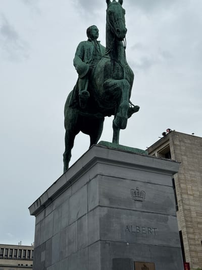 The image shows an equestrian statue of Albert I of Belgium, located in Place Poelaert, Brussels, Belgium. The statue is made of bronze and depicts Albert I in military attire, riding a horse. The statue stands on a granite pedestal which has the name "ALBERT" carved into it, along with a crown emblem. In the background, a modern building with stone facade and windows is visible, along with a section of another building with repeating vertical elements. The sky is overcast with clouds.