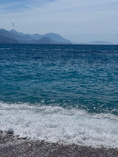 The image depicts a serene coastal scene with the Adriatic Sea in the foreground and a mountain range in the background. The water is a vibrant blue, with small waves gently lapping onto a dark, pebbly shore. In the distance, several layers of mountains fade into a hazy blue, suggesting a considerable distance. The sky is a pale blue with some wispy clouds. The overall impression is one of tranquility and natural beauty. This view is consistent with the landscape found in Prijevor, Montenegro, a region known for its stunning coastline and mountainous terrain. No people or specific structures are visible in the image.