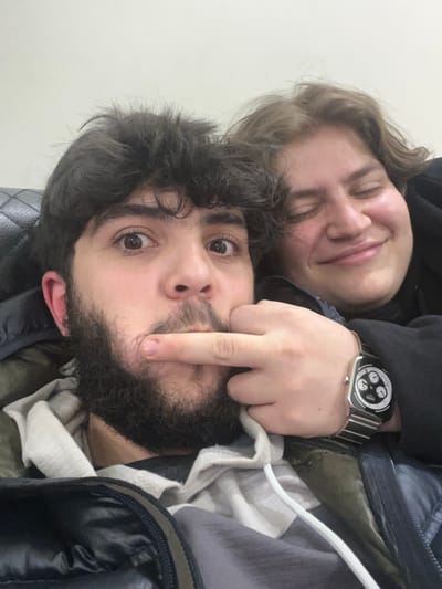 A selfie shows two people, a person with medium brown hair and closed eyes laying on top of a person with dark hair and a beard. The person with the beard is staring intently at the camera and has a finger raised towards their own face. The person on top has a black hoodie. The person on the bottom has a watch on their wrist and what appears to be a black puffer jacket. The image is rotated counter-clockwise, causing the background to appear to be a wall or floor.