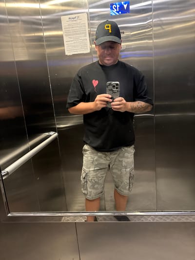 A man is standing in an elevator, taking a selfie. He is wearing a black t-shirt with a red heart graphic, camouflage cargo shorts, and a black baseball cap with a yellow "P" logo. He is holding a smartphone with a camouflage case. The elevator walls are made of polished stainless steel, and there is a handrail on the left side. A notice is attached to the wall to the left of the man. An illuminated sign with a blue background and white arrows is visible at the top of the elevator. The floor of the elevator has a diamond plate pattern.